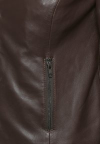 Nahaufnahme einer braunen Lederjacke mit einem schwarzen Reißverschluss auf der Vorderseite, glatter Textur und subtiler Geschmeidigkeit. Auffälliges Nähdetail.