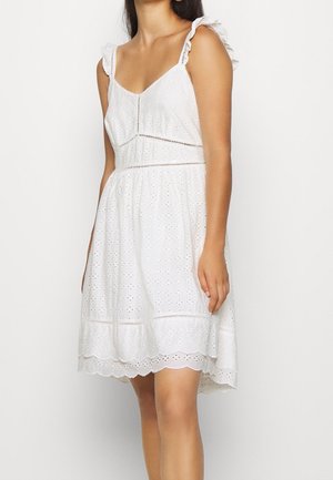 Kobieta w białej, bezrękawnikowej sukience typu eyelet z falbankowym dołem i wiązanymi ramiączkami, stojąca na jasnym, jednolitym tle.