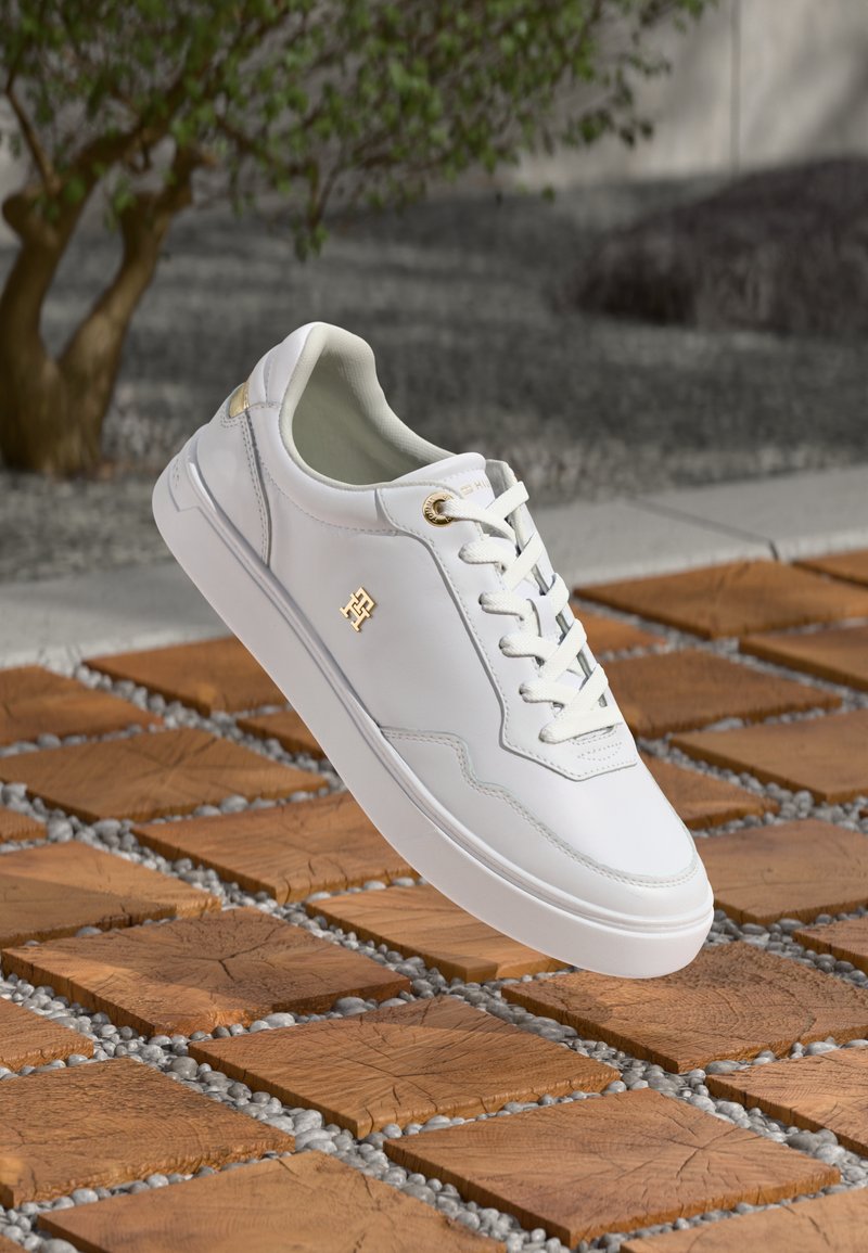 Sneaker bianco in pelle con dettagli dorati e lacci che fluttua sopra un pavimento in piastrelle di legno all'aperto, vicino a un albero.