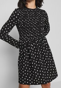 Czarna sukienka z długimi rękawami, z marszczonym materiałem w górnej części i białymi kropkami. Tkanina ma teksturowany wygląd, delikatnie rozszerzająca się w talii.