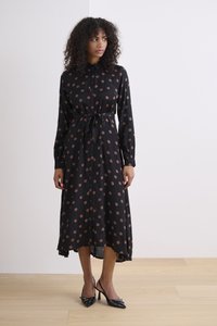 Schwarzes Kleid mit großen braunen Punkten, Knopfleiste, tailliertem Gürtel, langen Ärmeln, fließendem Saum und kombiniert mit schwarzen High Heels.