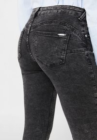 Jeans de denim negro con una cintura alta, diseño de cinco bolsillos y una textura de desvanecimiento sutil. La parte trasera tiene una costura curva y un pequeño acento metálico.