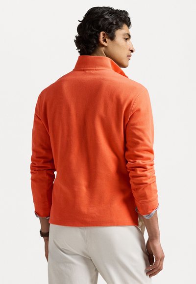 Pulover portocalie cu guler înalt, mâneci lungi și textură stil rib. Asortat cu pantaloni de culoare deschisă, văzut din spate.