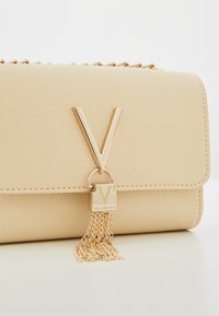 Béžová texturovaná kabelka Valentino se zlatým logem "V" a řetízkovou třásní na bílé ploše.