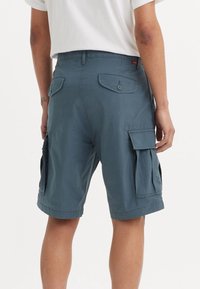 Osoba v modrých cargo šortkách s knoflíkovými zadními a bočními kapsami, oblečená v bílém tričku s krátkým rukávem, zachycena zezadu.