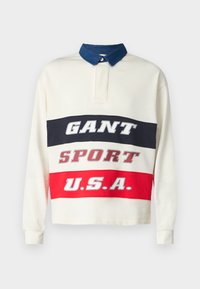 Langermet polo skjorte i krem med blå krage, med iøynefallende grafiske striper i marineblå, rød og hvit med tekst: "GANT," "SPORT," og "U.S.A."