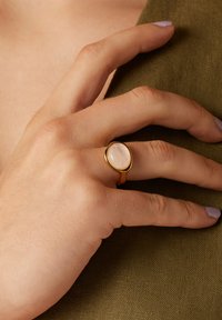 Gouden ring met een ovale rozenkwarts steen, met een glad oppervlak en een dunne band, gedragen op een hand tegen een textuur van olijfgroen stof.
