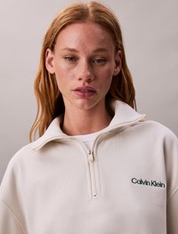 Bijela dukserica na zip s visokim ovratnikom, s zelenim izvezenim Calvin Klein logom, izrađena od mekog, teksturiranog materijala.
