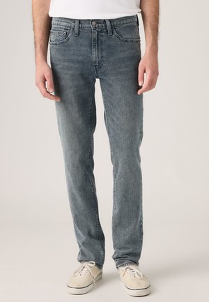 Homme portant un jean bleu délavé à jambes droites, des baskets blanches et une chemise blanche, debout devant un fond uni clair.