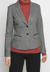 Grå houndstooth blazer med sorte detaljer, enkelt knaplukning, to forlommer og en skræddersyet pasform, lagd over en rød turtleneck.