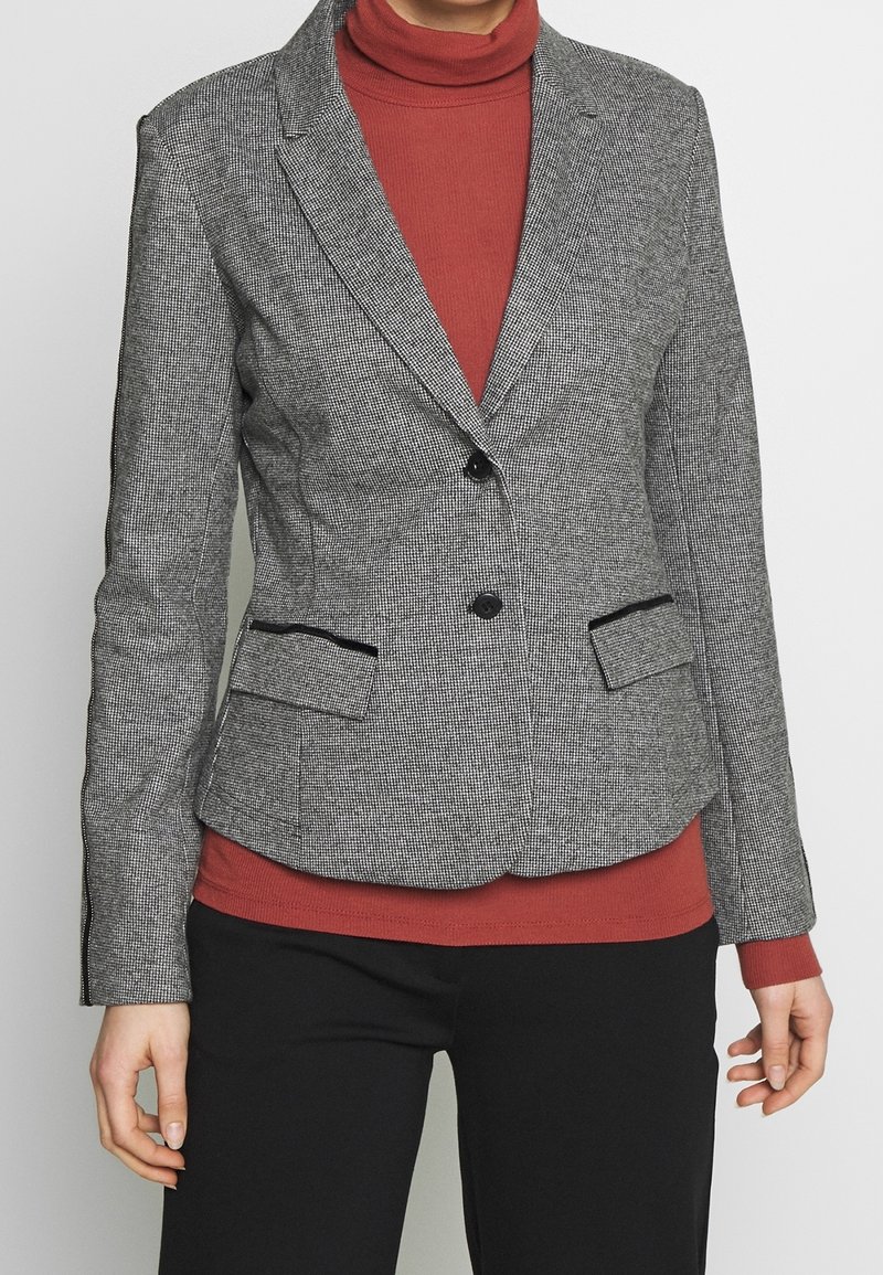 Grå houndstooth blazer med sorte detaljer, enkelt knaplukning, to forlommer og en skræddersyet pasform, lagd over en rød turtleneck.