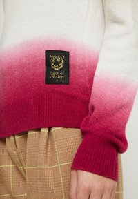 Gradientstickad tröja som övergår från vitt till rosa, med ribbade ärmar i en djup magenta. Har en svart etikett med guldbroderi.