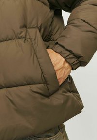 Doudoune en vert olive, avec un design matelassé, des poignets élastiques et des poches latérales. Le matériau semble lisse et léger.