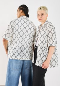 Weiße Kurzarmhemden aus Spitze mit schwarzen Rautenmuster, getragen von zwei Personen, kombiniert mit blauen und schwarzen Hosen.