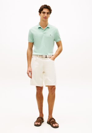 Világoszöld galléros póló, fehér rövidnadrág és barna szandálok. A pólón kis logó látható; a rövidnadrág laza szabású, zsebekkel.