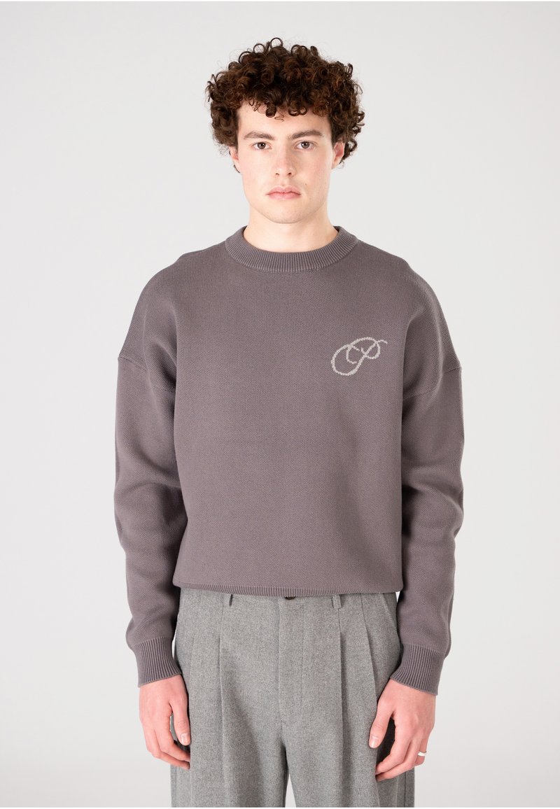 Pull court gris avec un tricot texturé, présentant un col rond et un logo brodé sur la poitrine. Assorti à un pantalon gris à plis.