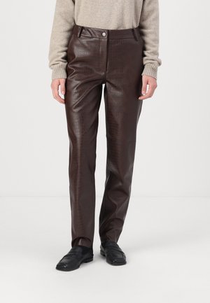 Personne portant un pantalon en cuir texturé brun foncé, un pull beige, des mocassins noirs, debout devant un fond clair uni.