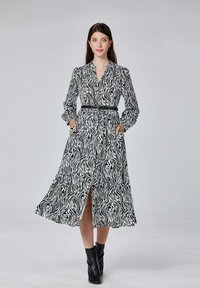 Robe midi imprimé zèbre noir et blanc avec des manches longues, un devant boutonné, une ceinture à la taille et des poches latérales. Associée à des bottines noires.