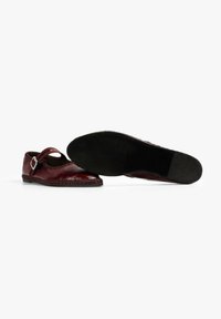 Zapatos de cuero burdeos con una correa y hebilla cuadrada, con la puntera redondeada y suela plana negra, texturizada con detalle trenzado a lo largo del borde.