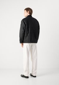 Black padded jacket with a collar, featured from the back. Worn with white pants and black shoes, showcasing a classic silhouette and texture.