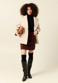 Beige kaksirivinen takki, musta ribattuna korkea kaulus, ruskea corduroy-hame, polviin asti ulottuvat mustat saappaat ja pieni ruskea käsilaukku.