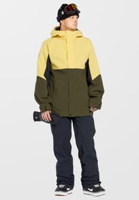 Veste imperméable jaune et verte avec capuche, associée à un pantalon de neige noir et des baskets noires. Tient un accessoire noir dans la main gauche.