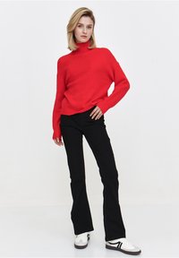 Czerwony sweter z golfem, robiony na drutach, z opuszczonymi ramionami, noszony z czarnymi dżinsami w kształcie dzwonów i białymi sneakersami z czarnymi akcentami.