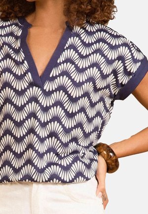 Vrouw draagt een marineblauw-wit patroon kortärmelig top met V-hals en witte broek, geaccentueerd met een bruine dikke armband.