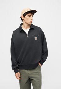 Ung mand iført beige kasket, sort kvart-zip sweatshirt med brystlomme og logo, samt olivengrønne bukser, ser til siden.