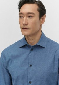 Jasnoniebieska koszula zapinana na guziki z wyraźnym kołnierzykiem, długimi rękawami i czarnymi guzikami. Tkanina ma subtelną teksturę.