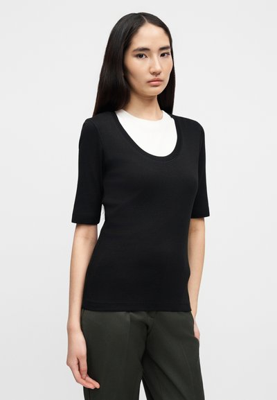 Μαύρο τοπ με κοντά μανίκια, φαρδιά ανοικτή λαιμόκοψη και εφαρμοστή γραμμή, κατασκευασμένο από λείο ύφασμα. Συνδυάζεται με πράσινο παντελόνι σε φαρδιά γραμμή.