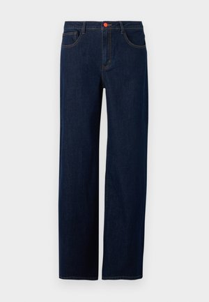 Persoon die donkere jeans met brede pijpen, zwarte Puma sneakers, een bruin overhemd en een roze vest met rits draagt, staand tegen een effen achtergrond.