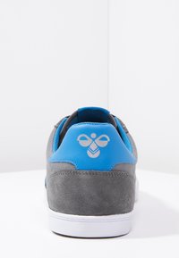 Grey suede sneaker with blue accents on the heel and tongue, featuring a white rubber sole and a logo displayed on the back.