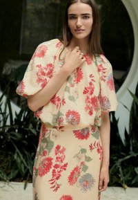 Blumenkleid in Creme mit lebhaften roten und blauen Mustern. Puffärmel, taillierter Schnitt, fließende Silhouette und strukturierter Stoff.