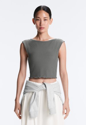 Femme face à l'objectif portant un crop top gris sans manches et un sweat-shirt gris clair noué autour de sa taille sur un fond uni.
