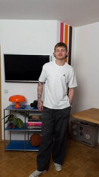 Νεαρός με τατουάζ, ντυμένος με λευκό πουκάμισο και σκούρο παντελόνι, στέκεται σε ένα δωμάτιο με μια μπάλα μπάσκετ, πορτοκαλί φωτιστικό, τηλεόραση και μια βαλίτσα με αυτοκόλλητα.