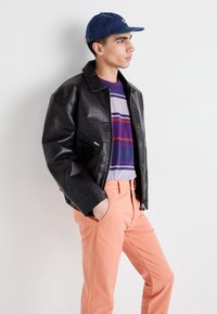 Black leather jacket, purple shirt with multi-colored stripes, and coral pants. Model wears a blue cap. Simple background.
