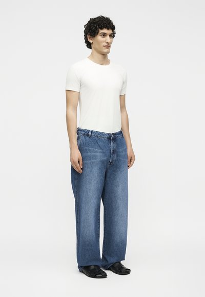 Άνδρας με σγουρά μαλλιά, ντυμένος με εφαρμοστή λευκή μπλούζα, φαρδιά μπλε τζιν και μαύρα σανδάλια, στέκεται μπροστά σε απλό λευκό φόντο.