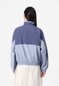 Žena s dlouhými tmavými vlasy, oblečená v dvoubarevném modrém větrovém kabátu s elastickými manžetami a spodním lemem, stojící zády k bílé pozadí.