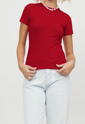 Person wearing a red short-sleeve shirt, light blue jeans, a pearl necklace, and a ring, holding the shirt hem with both hands.