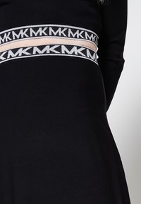 Haut noir à manches longues avec une coupe ajustée, doté d'une large ceinture décorée d'un motif répétitif "MK" en blanc.