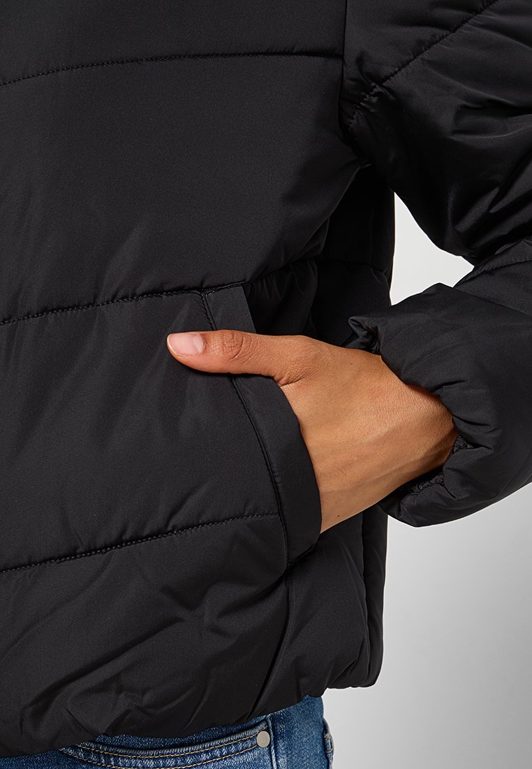 Mano nella tasca di un giubbotto nero imbottito con polsini in elastico, che mostra i dettagli della cucitura e una parte dei jeans in denim blu.