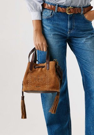 Person trägt blaue Jeans und gestreiftes Hemd und hält eine braune Wildleder-Handtasche mit dunklen Griffen und Quasten seitlich.