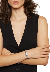 Silver braided bracelet with a smooth, rectangular clasp, worn on a bare wrist. The background features a black, sleeveless outfit.