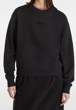 Person iført en sort Vans-sweatshirt med lange ærmer og ribbet kant, kombineret med sorte bukser.