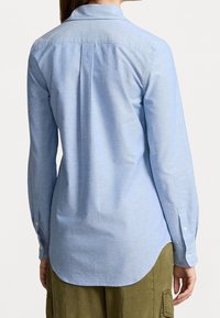 Ljusblå långärmad skjorta med en bakfåll, böjd nederkant och knappade manschetter. Tyget verkar slätt och något strävt.