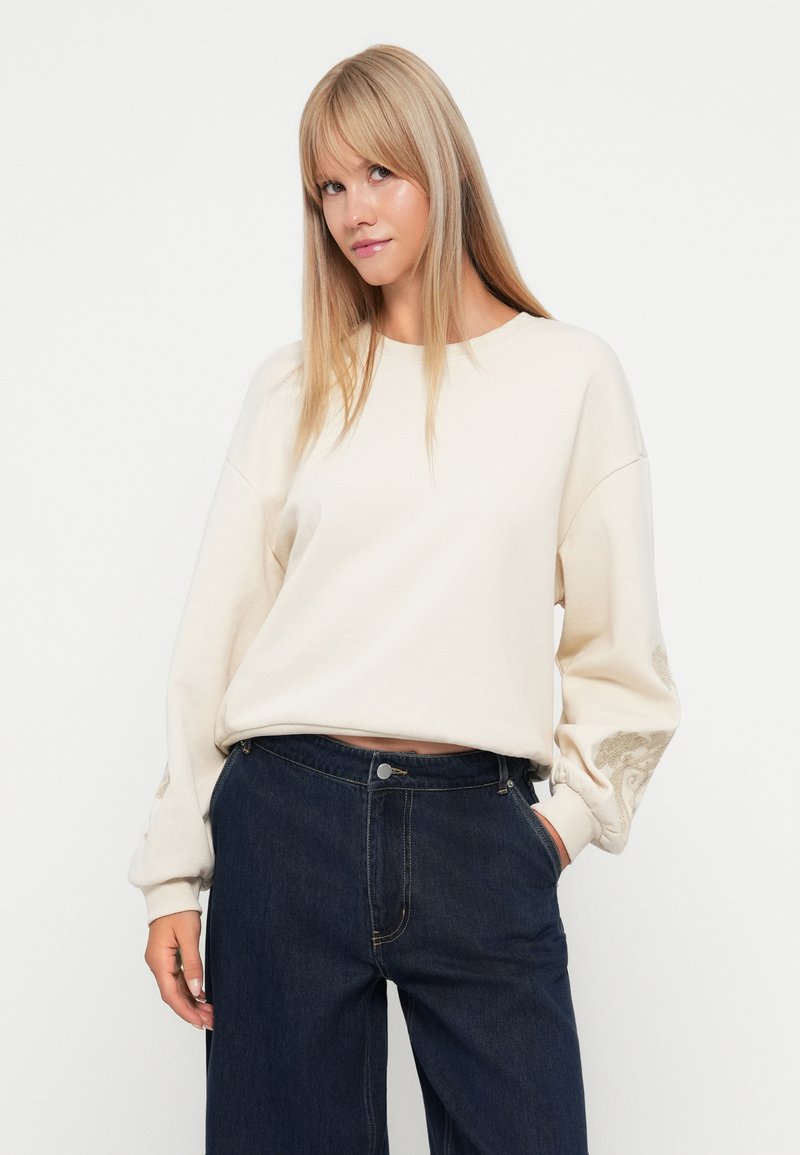 Ivory sweatshirt z okrągłym dekoltem i luźnym krojem. Posiada haftowane detale na rękawach. Noszony z ciemnoniebieskimi spodniami o szerokich nogawkach.