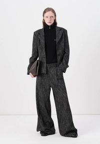 Černý kostičkovaný blazer s širokými klopami, kombinovaný s přiléhavým černým rolákem. Volné černé kostičkované kalhoty a hnědá kabelka na ruku.