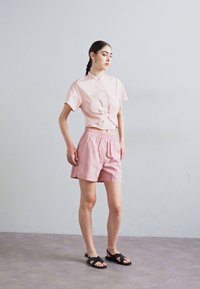 Camicia rosa chiaro a maniche corte con dettaglio anteriore annodato. Pantaloni corti a righe rosa chiaro con cintura elastica. Sandali neri con suola piatta.