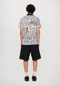 Camisa de manga curta com padrões de texto manuscrito em preto e branco, com colarinho, combinada com calções pretos sólidos e sapatos pretos.